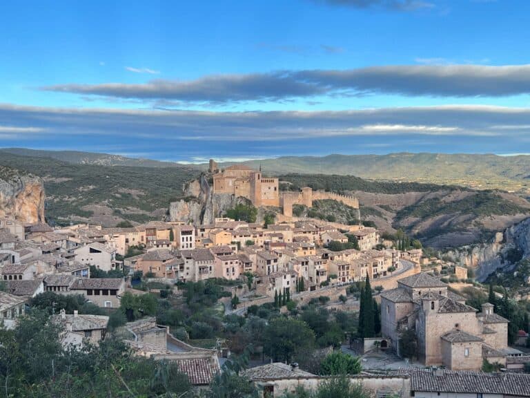 Panoramic view of Alquezar Spain at sunset from a lookout point on the outskirts of town, showing the stone buildings and the castle against the backdrop of the rolling green hills and darkening blue sky.