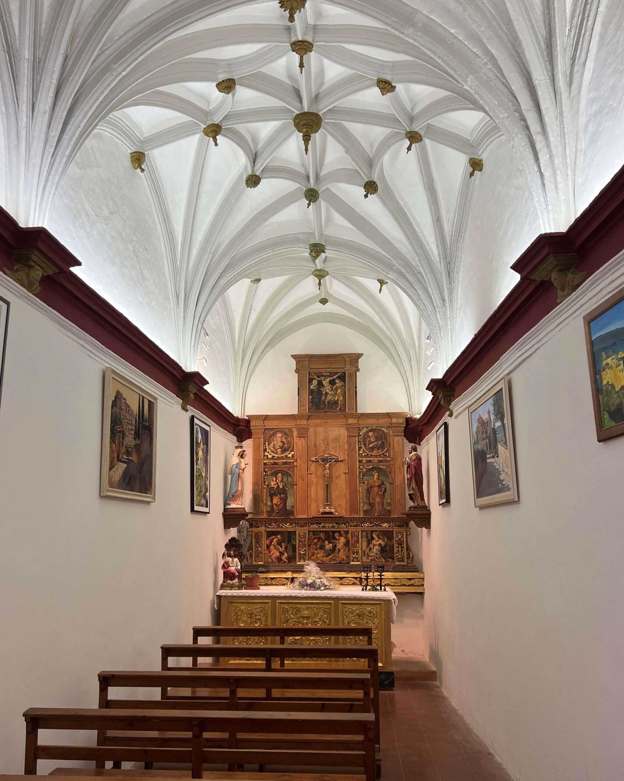 Inside a small chapel in Alquezar with intricate arched white ceiling, wooden benches, and the altar with the Jesus on the cross. 