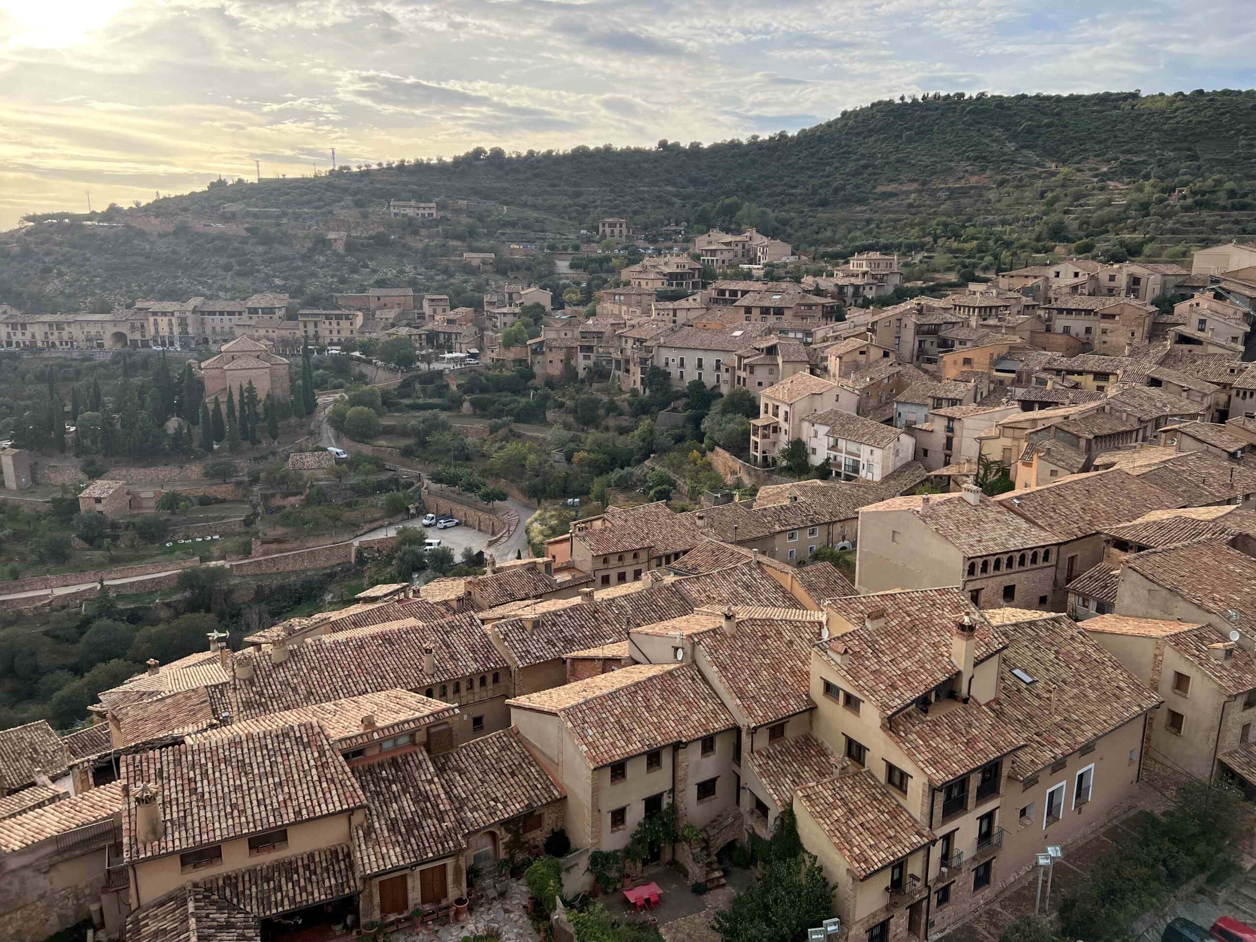 View from above the small village of Alquezar Spain. Small tan buildings built into the rolling green and rocky hillside with the sun setting in the background.