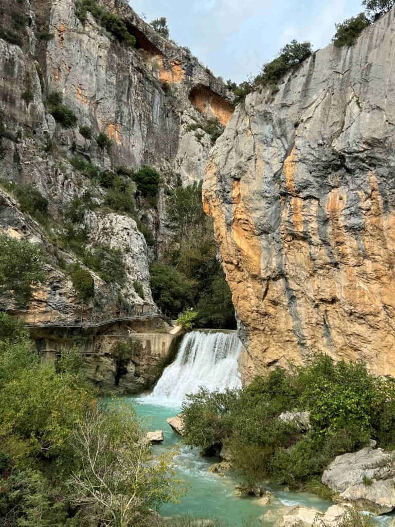 View from inside a rocky canyon with a short waterfall running into a turquoise blue river and greenery surrounding the river in the forefront.