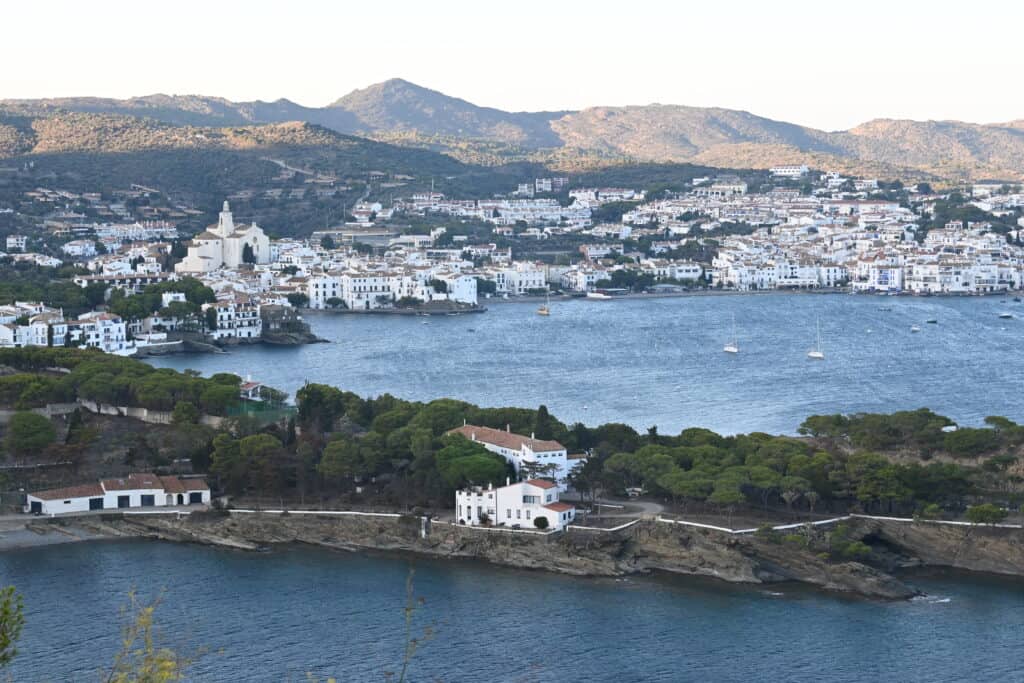 Far away shot of the town of Cadaques with a cluster of the white buildings in town lining the blue Mediterranean shoreline.