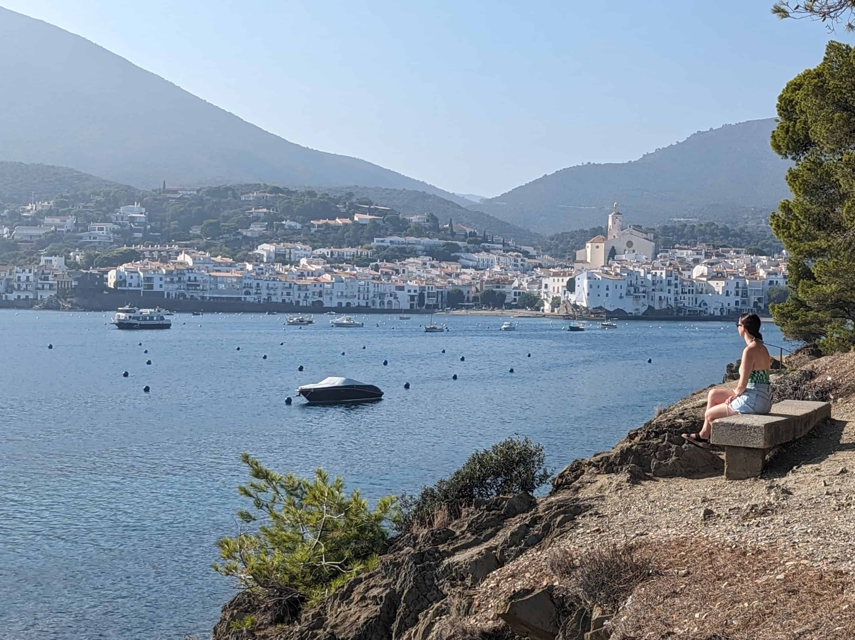 Girl sitting on a stone bench overlooking the Mediterranean Sea and the town of Cadaques, Spain with small white buildings on the shoreline.