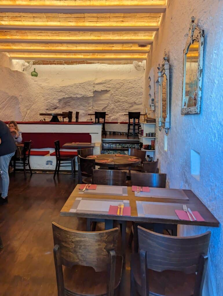 Interior of a small restaurant with a handful of empty wooden tables against white washed stone walls.