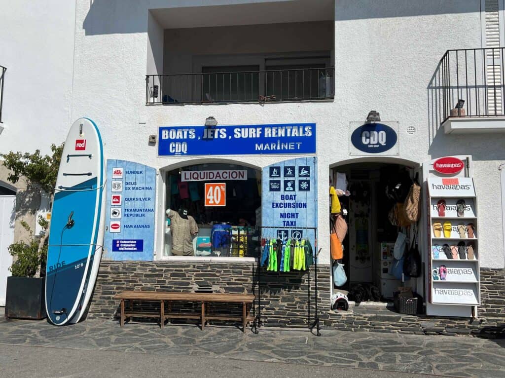 A boat and water sport rental shop storefront in Cadaques. Surfboards, a bench, and scuba flippers sit out front on the street.