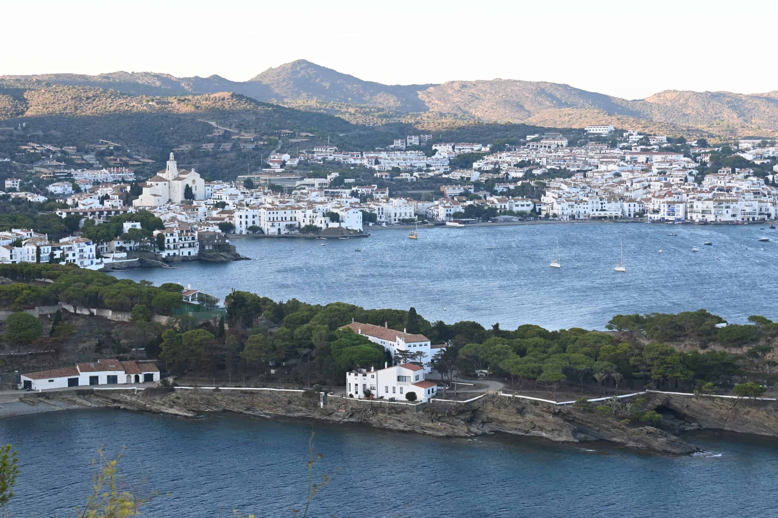 Far away shot of the town of Cadaques with a cluster of the white buildings in town lining the blue Mediterranean shoreline.