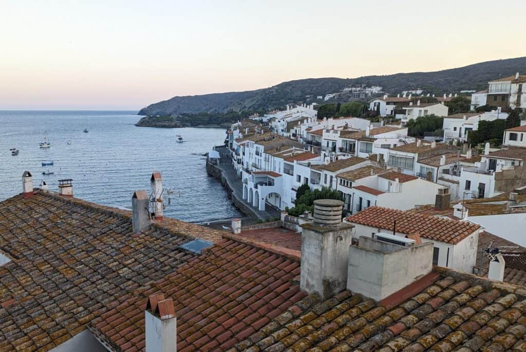 View of Cadaques at sunset with terracotta roofs on small white buildings lining the coastline.