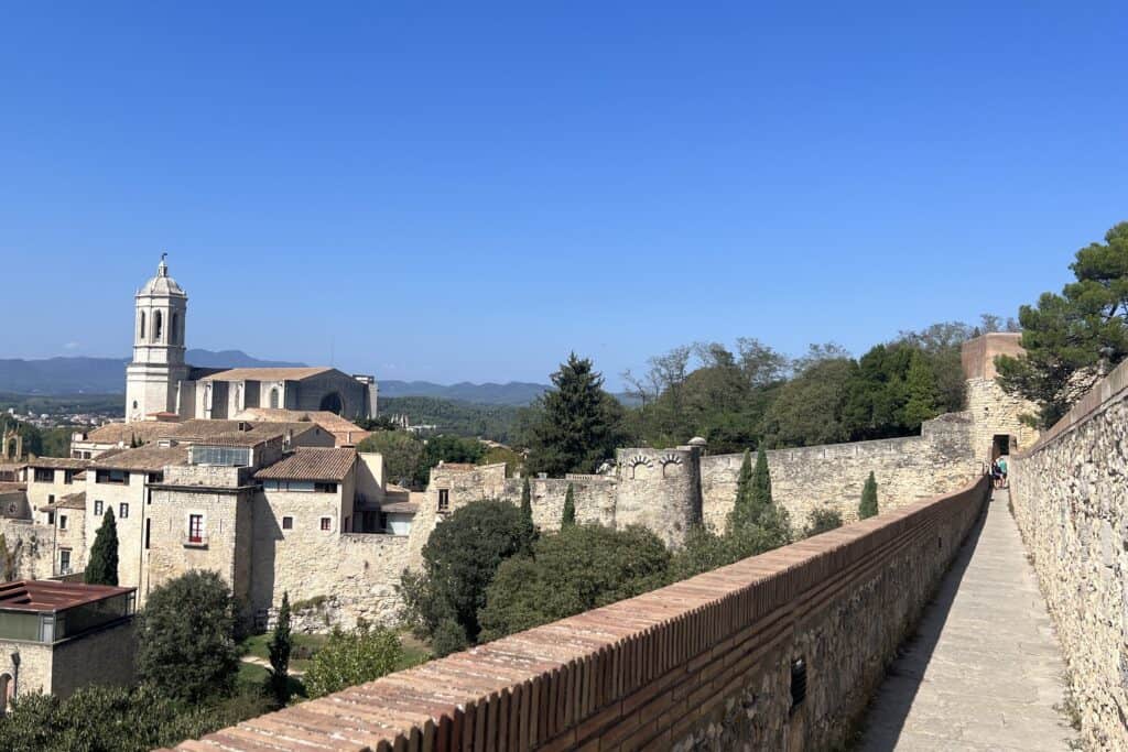 View from walking on the City Walls in Girona, Spain including the stone wall path on the right and the Cathedral on the left with trees and blue sky surrounding both structures.