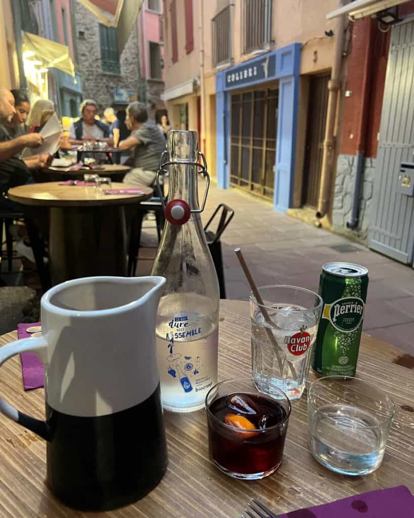 Small wooden table with a carafe, a glass of sangria, and other clear beverages on the street outside a restaurant in Collioure.