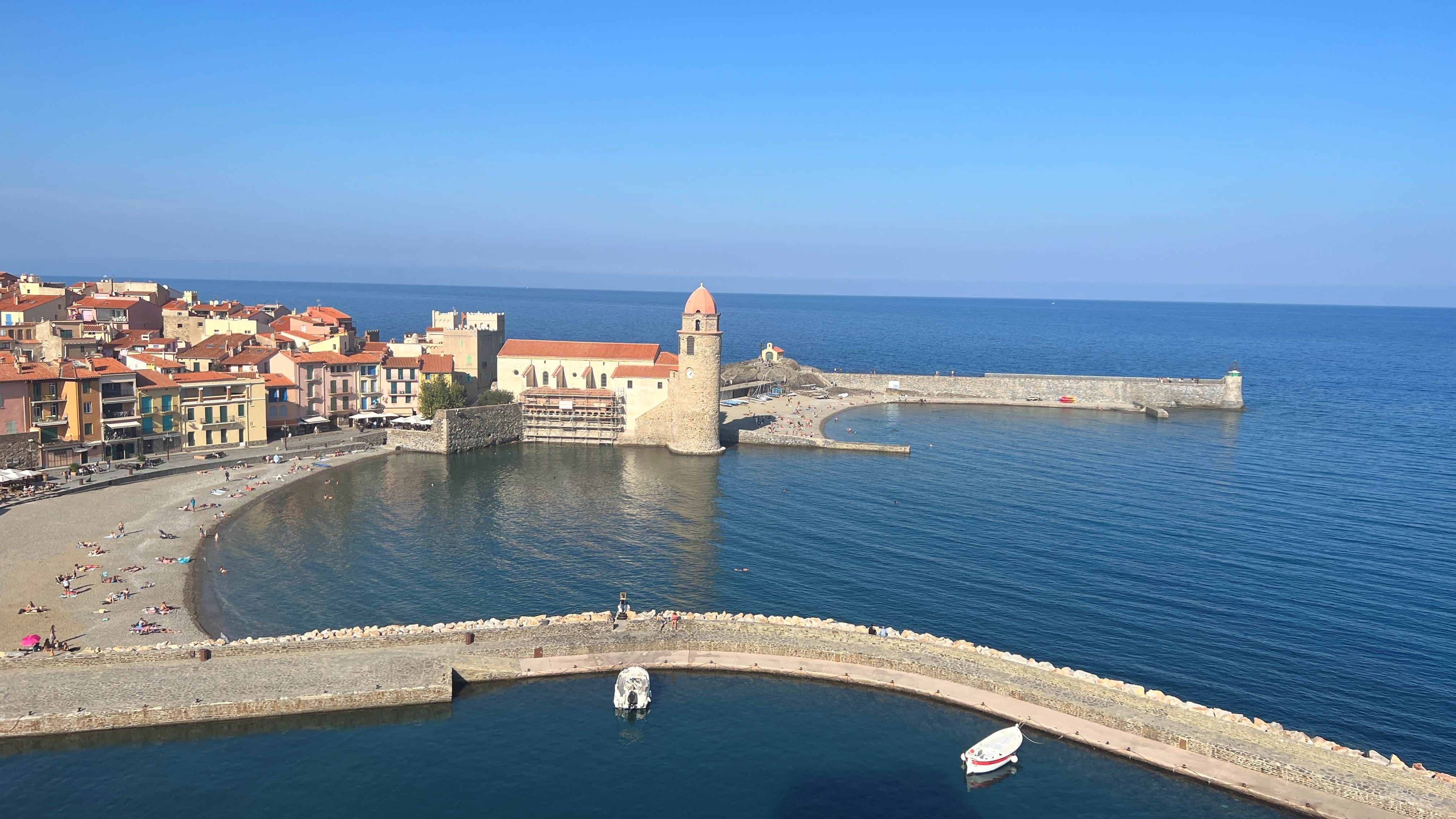 View of Collioure from the castle which shows a pebble beach, lighthouse and sea wall extending out into the blue sea.