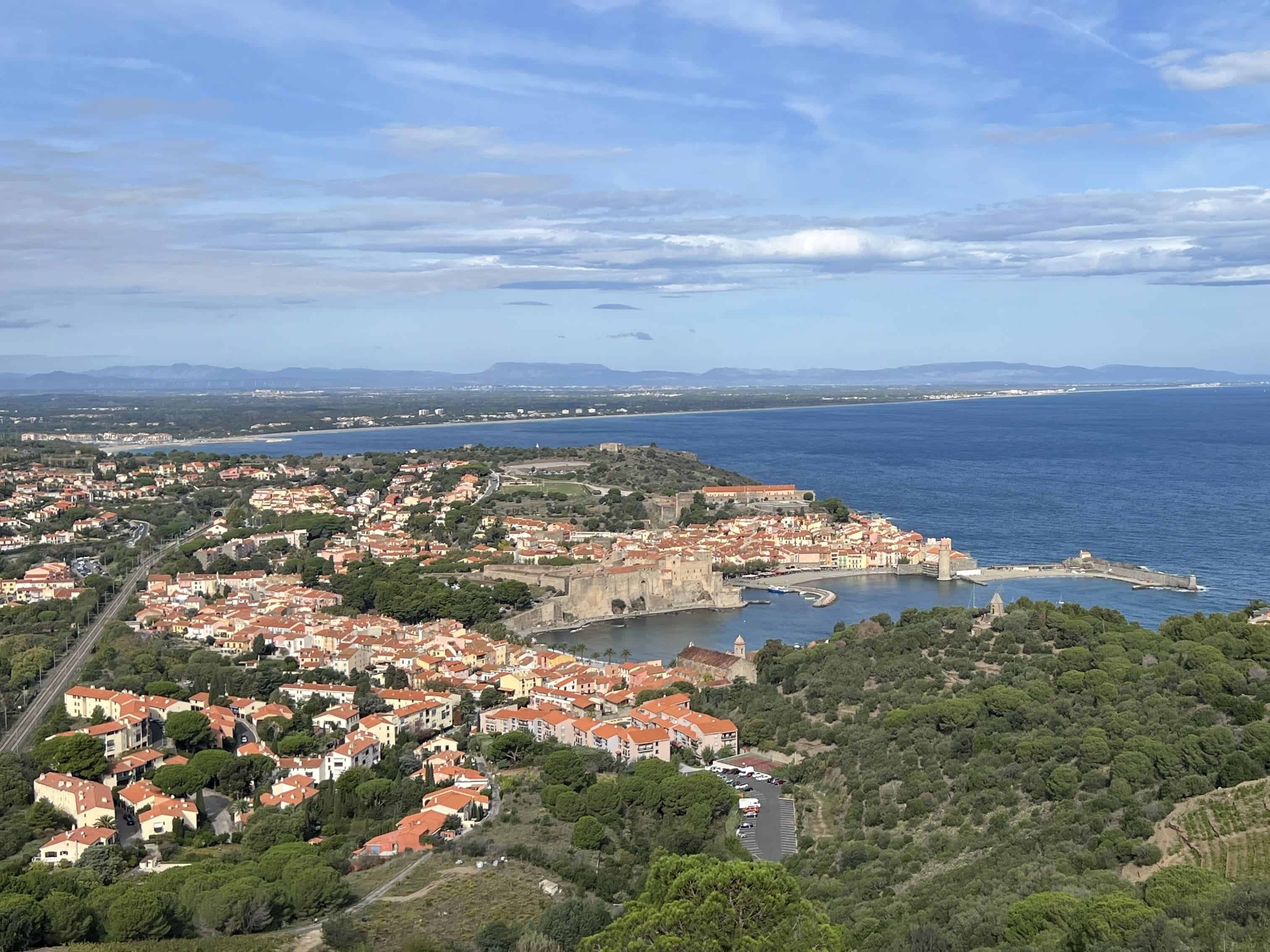 View from the hillside above Collioure France which shows the small light brown buildings along the green coastline with the blue sea on the right and lightly cloudy sky above.