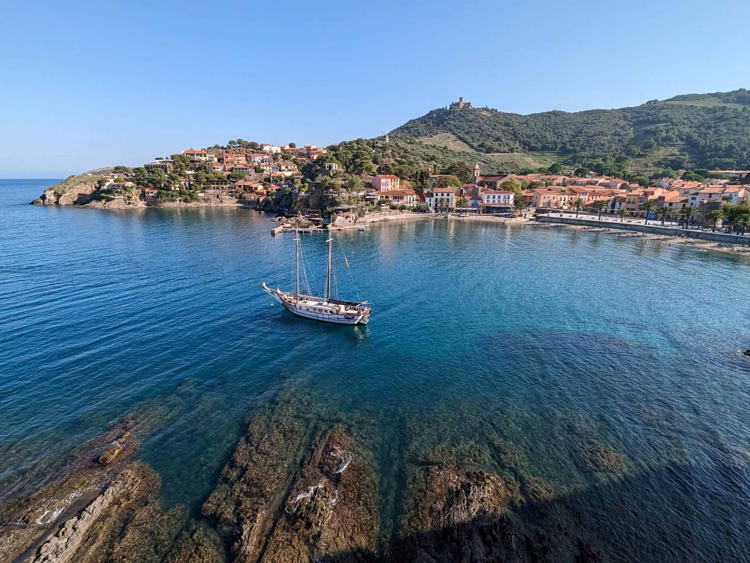 View of the Mediterranean Sea from the castle in Collioure, France. Clear blue water with a boat with small buildings lining the hilly shore in the background.