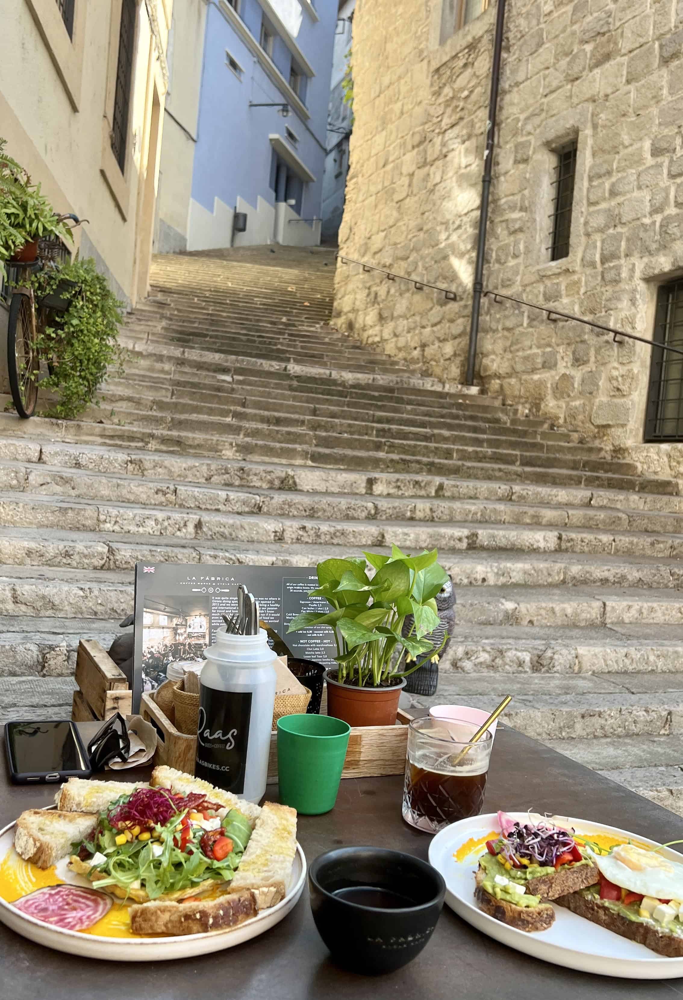 Two breakfast dishes with avocado toast on a small black table with a stone staircase in the background, outside of La Fabrica, a cafe in Girona Spain.