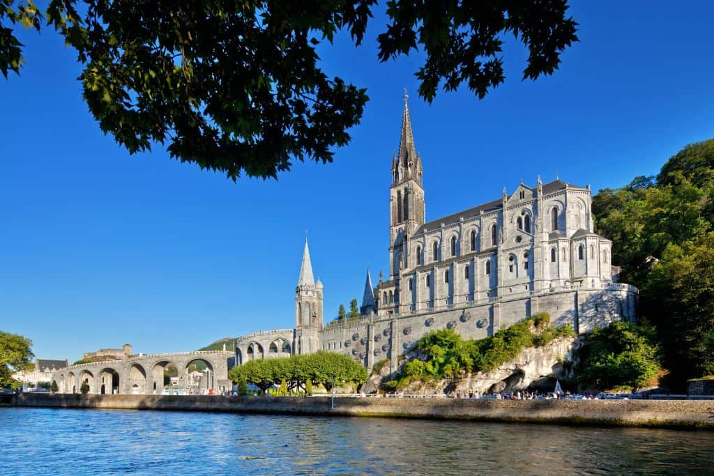 A large, ornate Catholic church in Lourdes overlooking the river. The church has a tall steeple and many windows. Trees and greenery surround the church. People are walking along the riverbank.