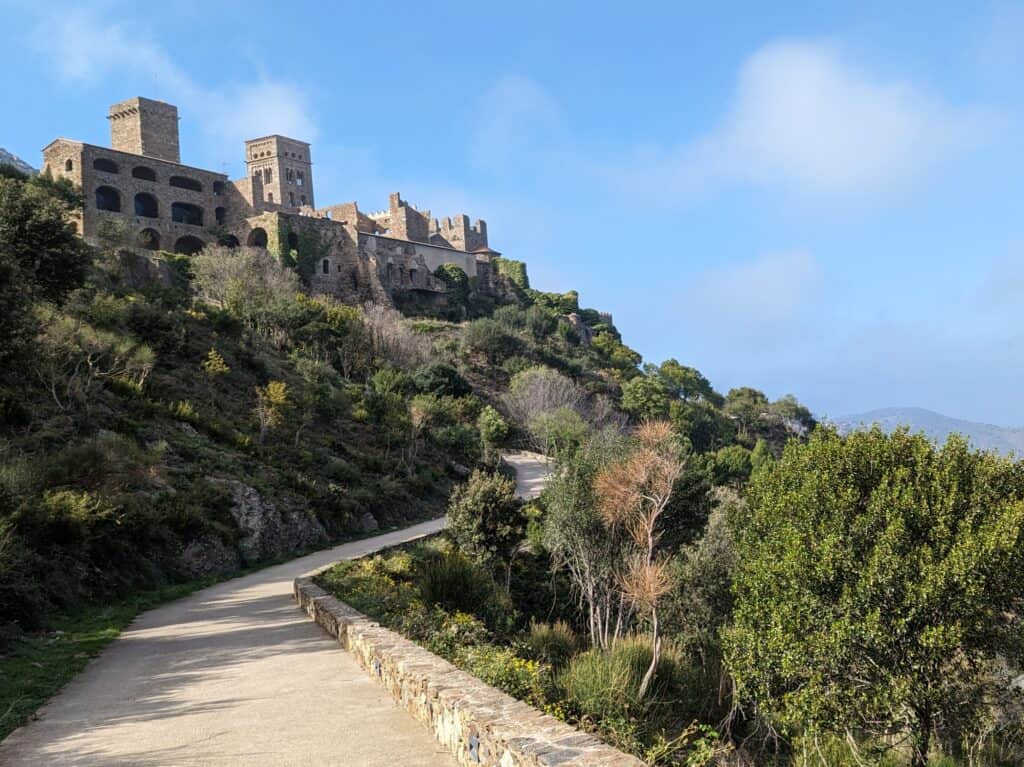 View of walking trail leading up to Monestir de Sant Pere de Rodes with the large stone monastery on the left and the green hillside on the right.