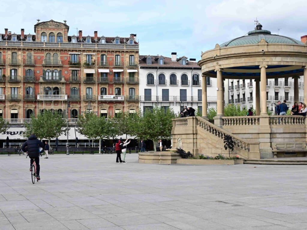 A Spanish square with a cafe called "Iruña." The cafe has a large, outdoor seating area with people enjoying drinks and food. There are buildings and trees in the background with a circular gazebo in the middle of the square.