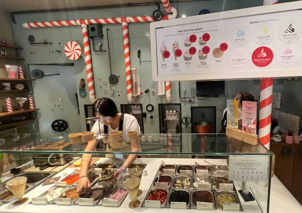 Ice cream shop with a person scooping ice cream from a counter with several flavors and toppings on display through a glass case.