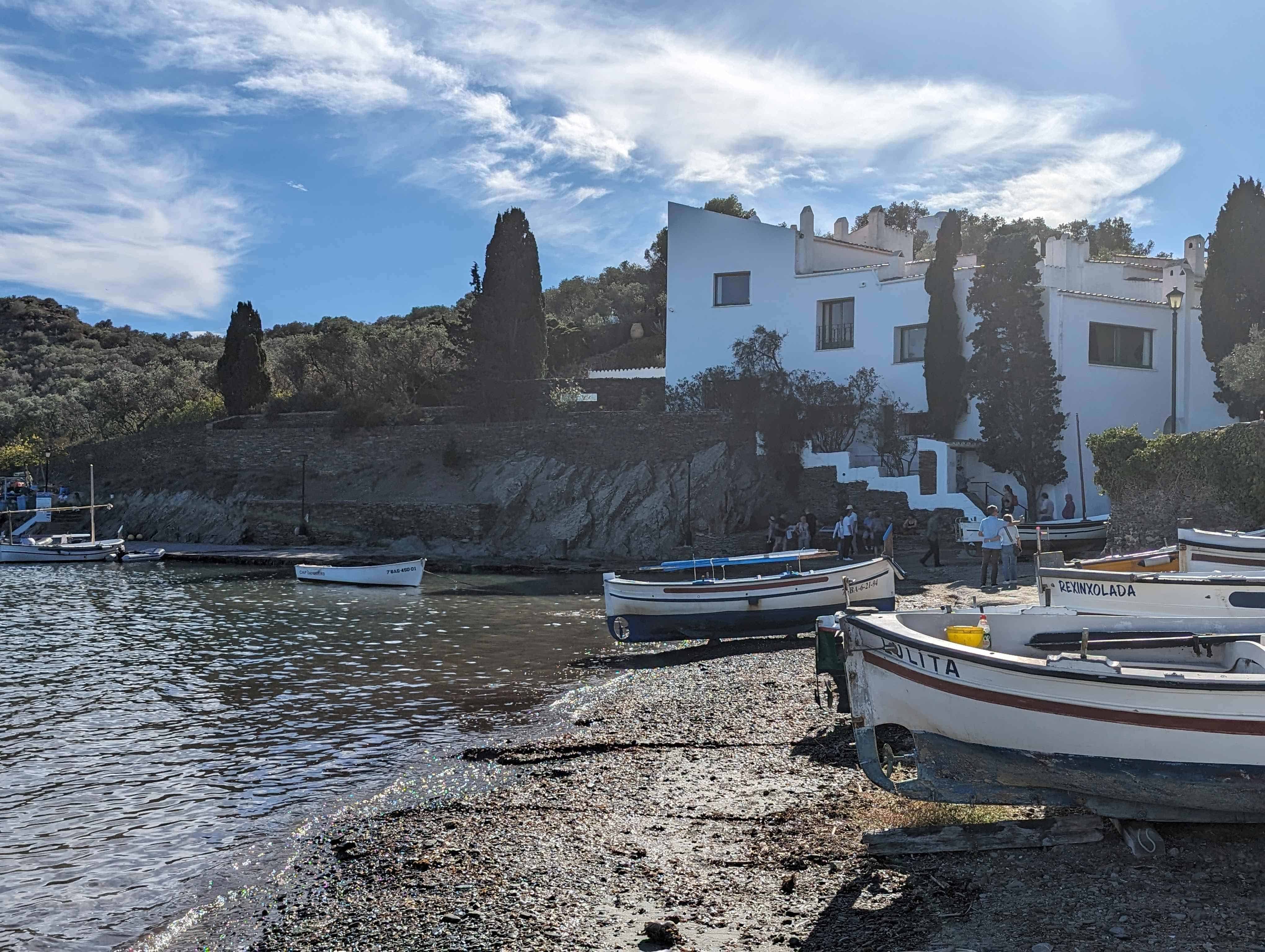 View of Salvador Dali's house in Port Lligat. The white house is now a museum that sits next to the water with small boats resting along the shoreline in front.