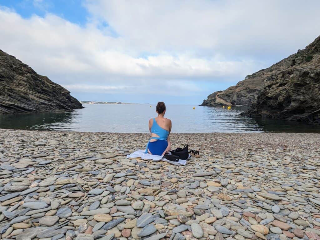 Girl in blue bathing suit sitting on a stone beach in a cove at Cap de Creus National Park.