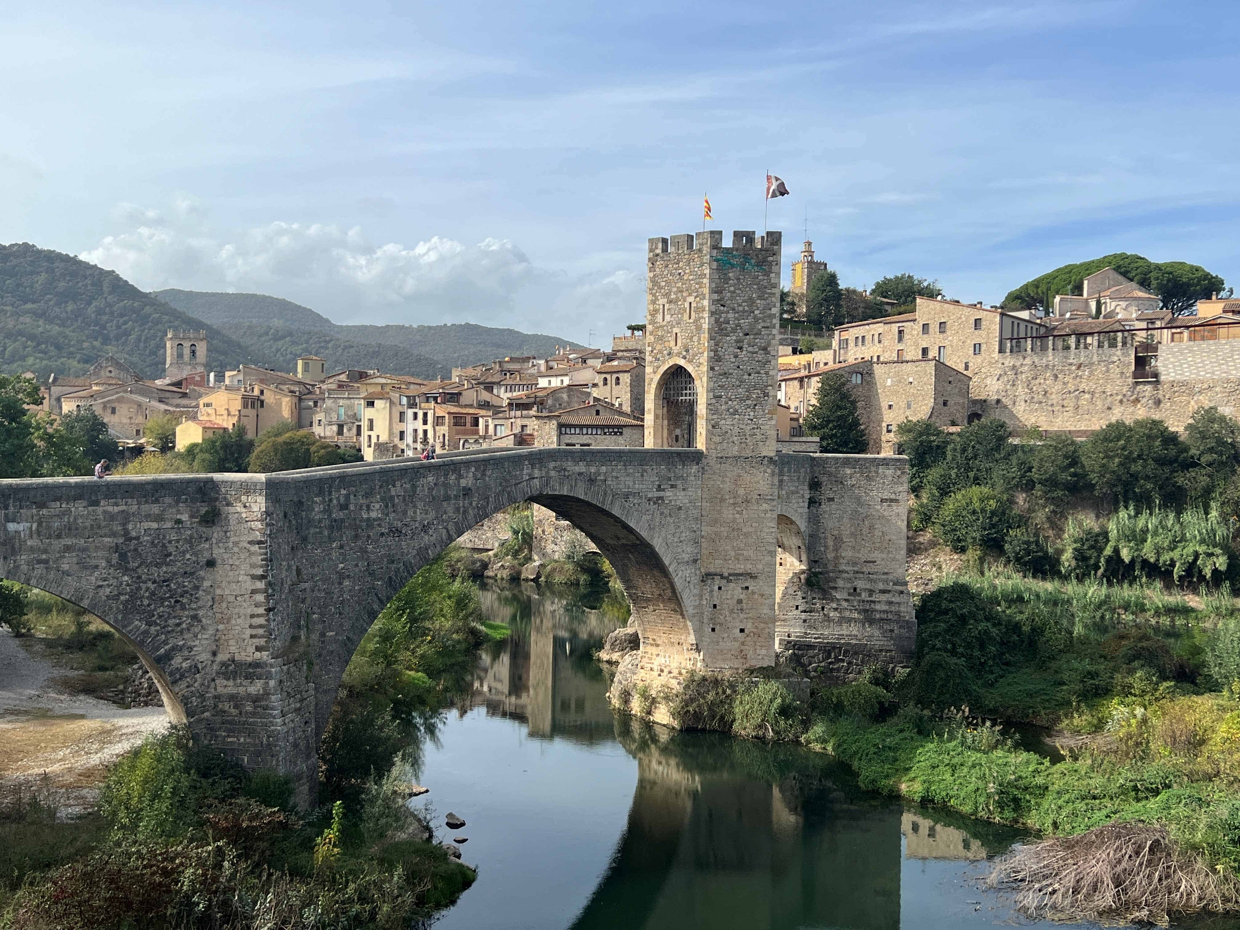 A medieval stone bridge in Besalú, Spain, spanning a calm river with reflections of the archway and nearby buildings. The town's historic architecture and hilly landscape are visible in the background under a partly cloudy sky.