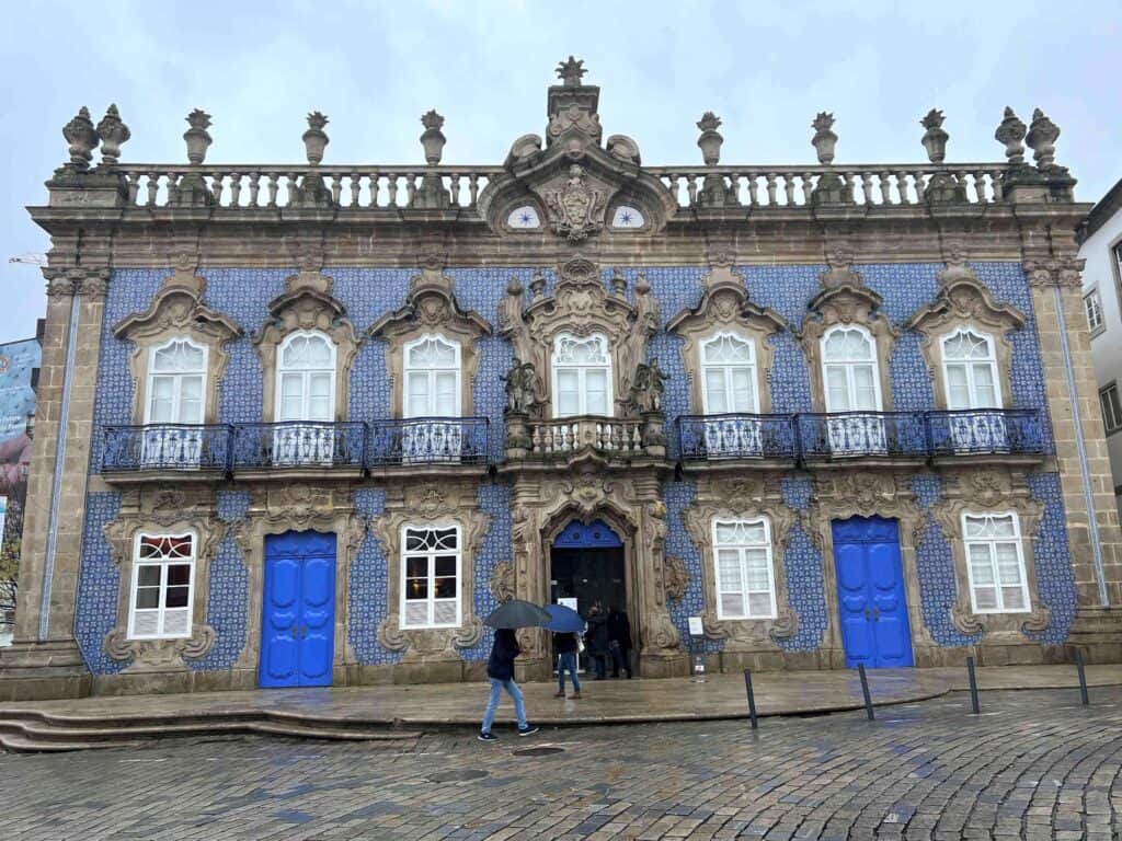 The Palácio do Raio, a historic Baroque building in Braga, Portugal, features a facade adorned with blue and white azulejos (ceramic tiles) and intricate stonework. Large, ornately framed windows and blue wrought-iron balconies line the building, while vibrant blue doors stand out against the detailed stone carvings. Statues flank the main entrance, adding to the grand appearance. Two people with umbrellas walk past on the rain-soaked cobblestone street.