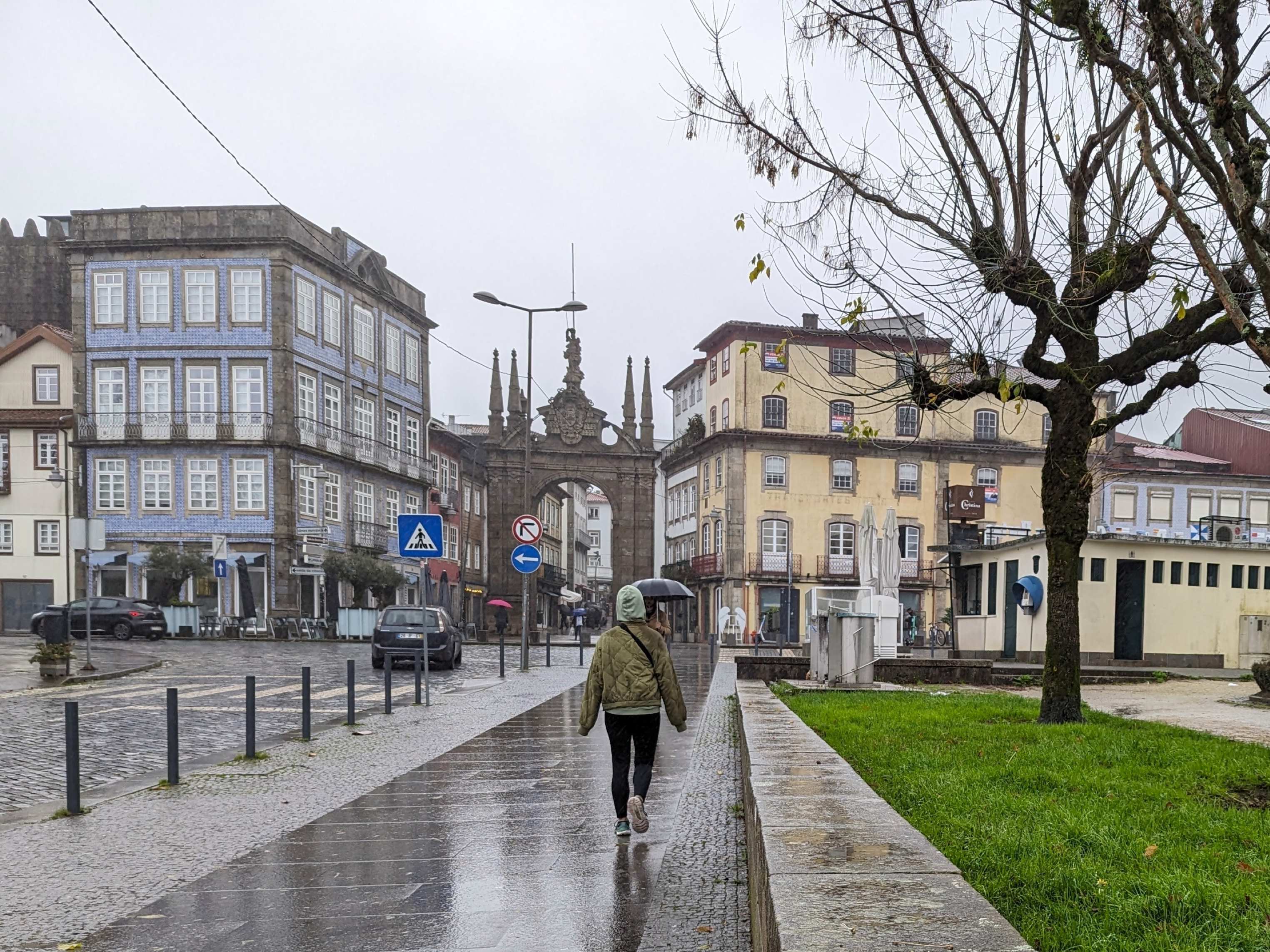 A rainy street scene in Braga, Portugal, with a person walking toward the historic Arco da Porta Nova, an ornate stone archway. Surrounding buildings feature classic European architecture, including a blue-tiled facade and yellow-painted walls. A leafless tree stands on the right, and reflections of the buildings shimmer on the wet pavement.