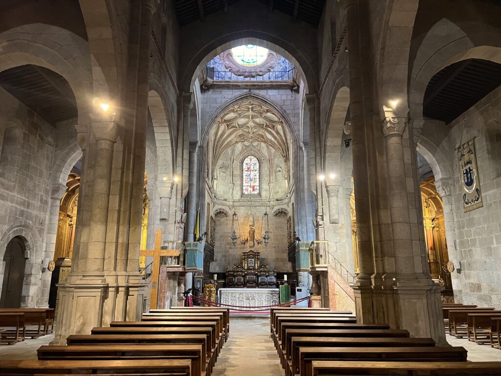 The interior of Braga Cathedral in Portugal, displaying a Romanesque and Gothic architectural style with tall stone arches and ribbed vaulted ceilings. Wooden pews line the central aisle, leading toward an intricately designed altar adorned with religious icons and a stained glass window above, casting soft light over the sanctuary. Decorative banners hang on the side walls, and the stone columns and walls give the space a historic and reverent atmosphere.