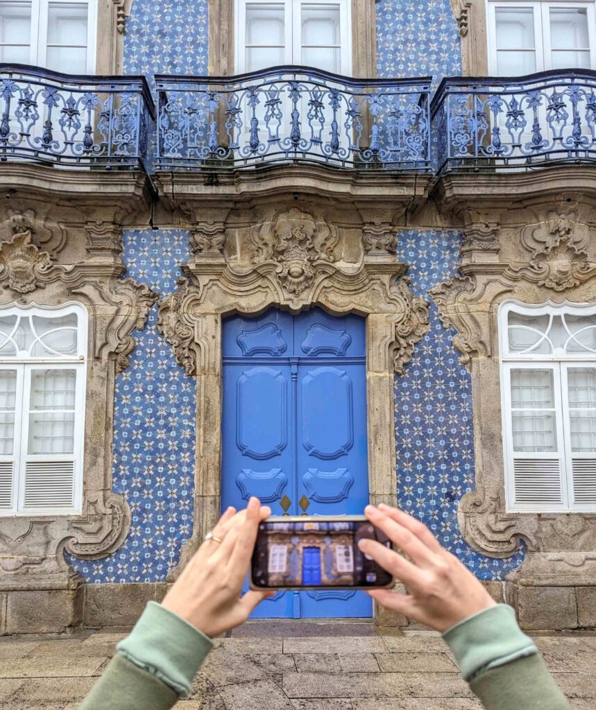 A grand, ornate building facade with intricate architectural details, including a large blue wooden door framed by elaborately carved stone elements. The building is adorned with a patterned tile wall and decorative wrought-iron balconies. A person's hands are shown holding a smartphone, capturing a photograph of Palacio do Raio.