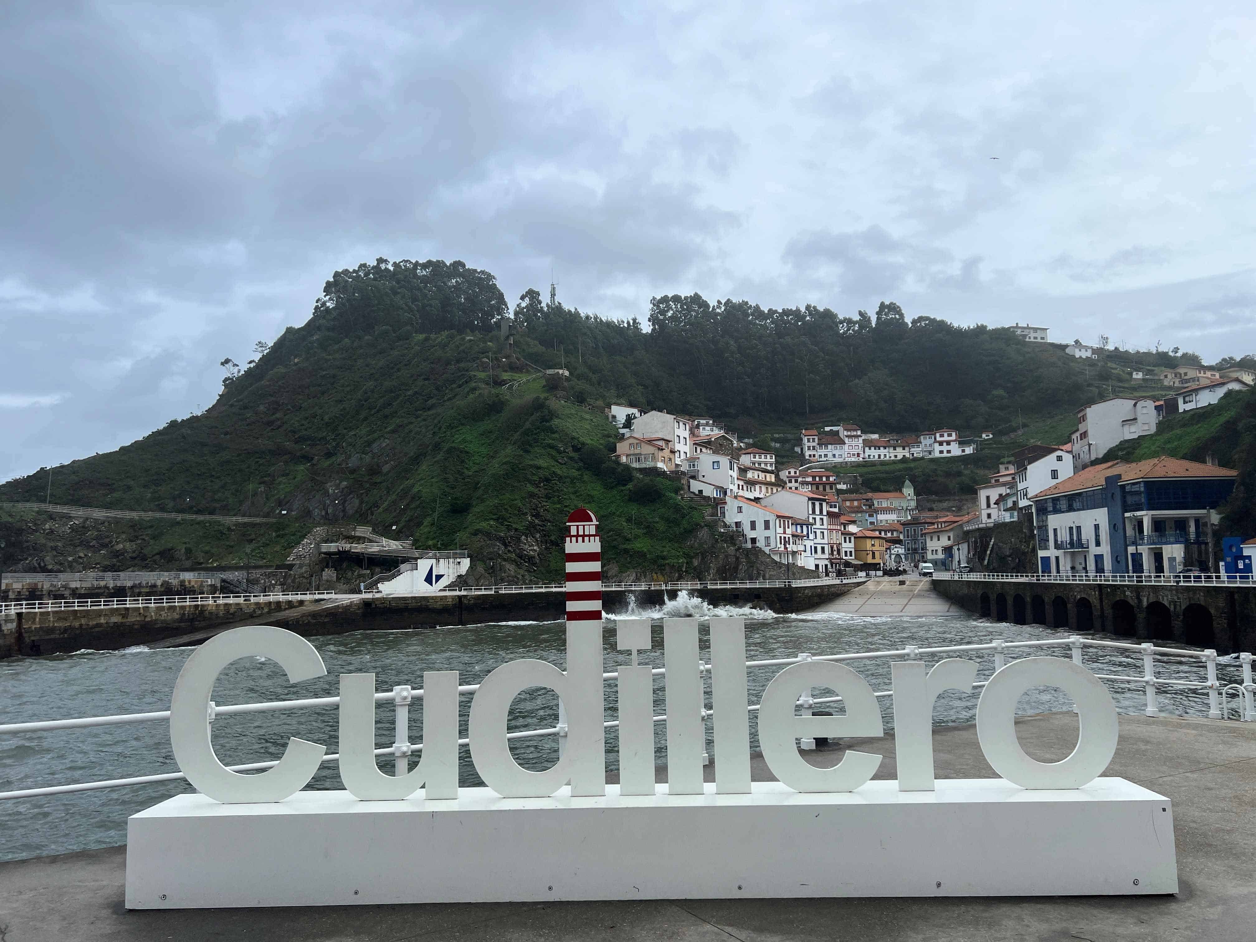 A coastal view of Cudillero, Spain, with a large white sign in the foreground spelling "Cudillero" and a red-striped lighthouse nearby. The hillside town with whitewashed buildings is visible in the background under a cloudy sky