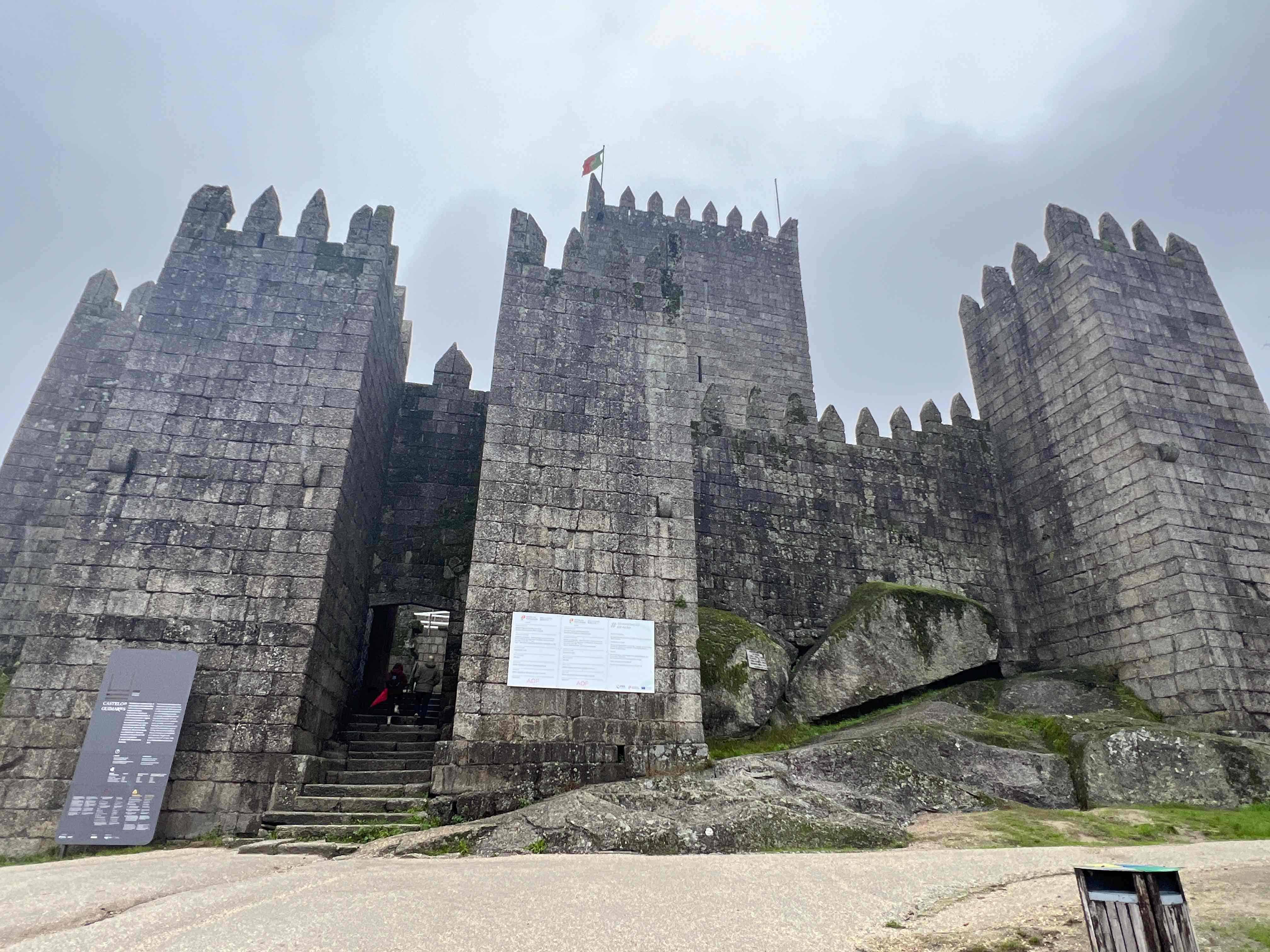 Stone fortress with tall, crenellated walls and an arched entrance, surrounded by large moss-covered rocks, under a cloudy sky. A Portuguese flag flies atop one of the towers of Guimaraes Castle.