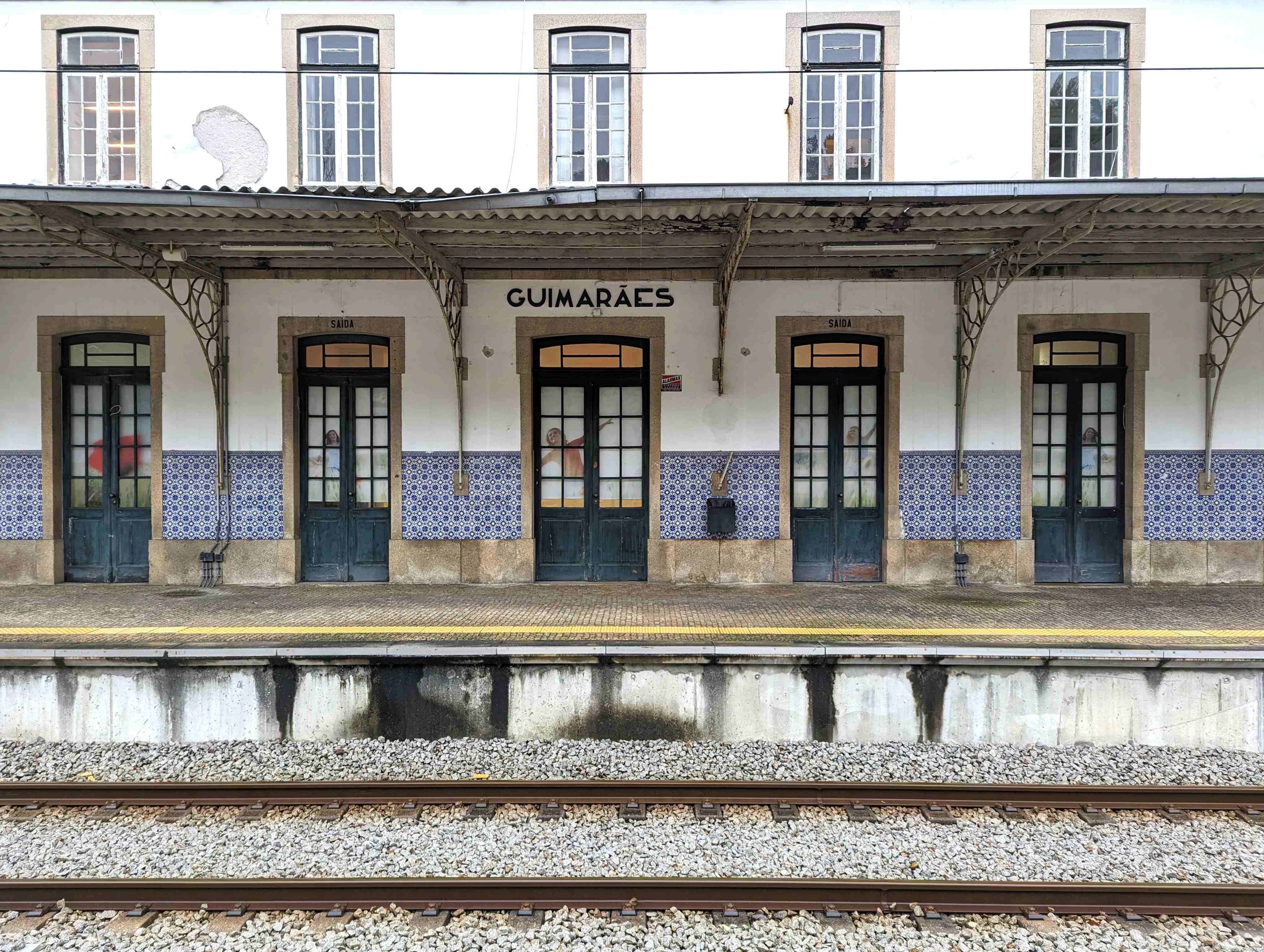  view of Guimarães train station in Portugal, featuring a row of tall, dark green doors with arched windows. The building’s facade is adorned with traditional blue and white Portuguese tiles along the lower half, with the station name 'Guimarães' displayed above the central door. Railroad tracks run across the foreground, and an overhanging metal roof with ornate supports provides shelter above the platform.