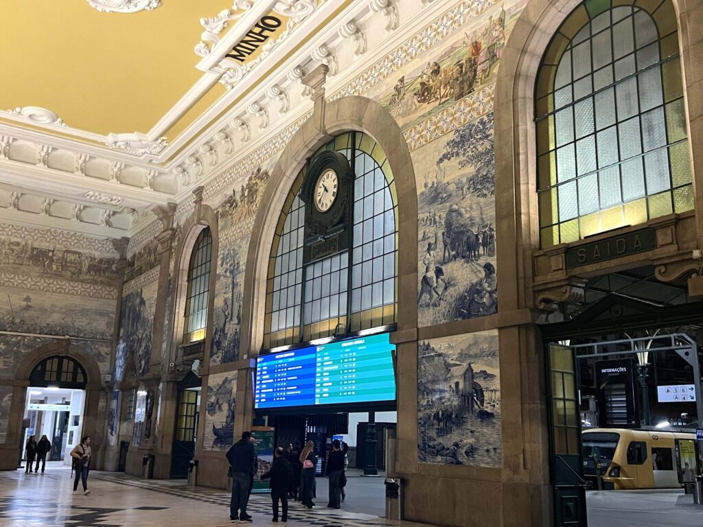 The interior of São Bento train station in Porto, Portugal, featuring grand arched windows and walls decorated with traditional blue and white azulejo tiles depicting historical scenes. The high ceiling has intricate moldings and a warm yellow tone. A large clock hangs above the main departure board, and several people stand in the spacious hall, looking at the timetable or waiting near the platform entrance. The scene captures the architectural elegance and cultural richness of the station.