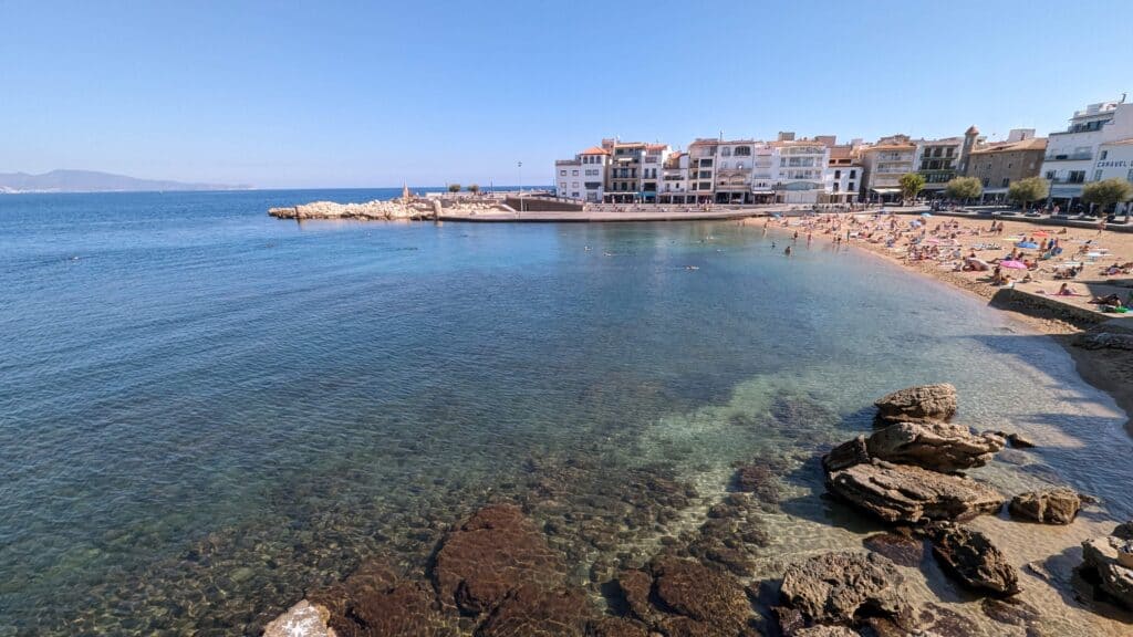 A view of a beach in L'Escala, Spain, featuring clear blue waters, rocky outcrops, and people sunbathing and swimming. The beach is lined with whitewashed buildings along the waterfront, under a clear blue sky.