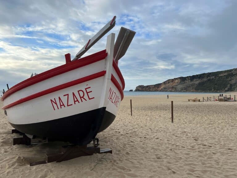 View of Nazare beach with a large wooden fishing boat perched on the sand on the left and the cliff side leading out to the ocean on the right.