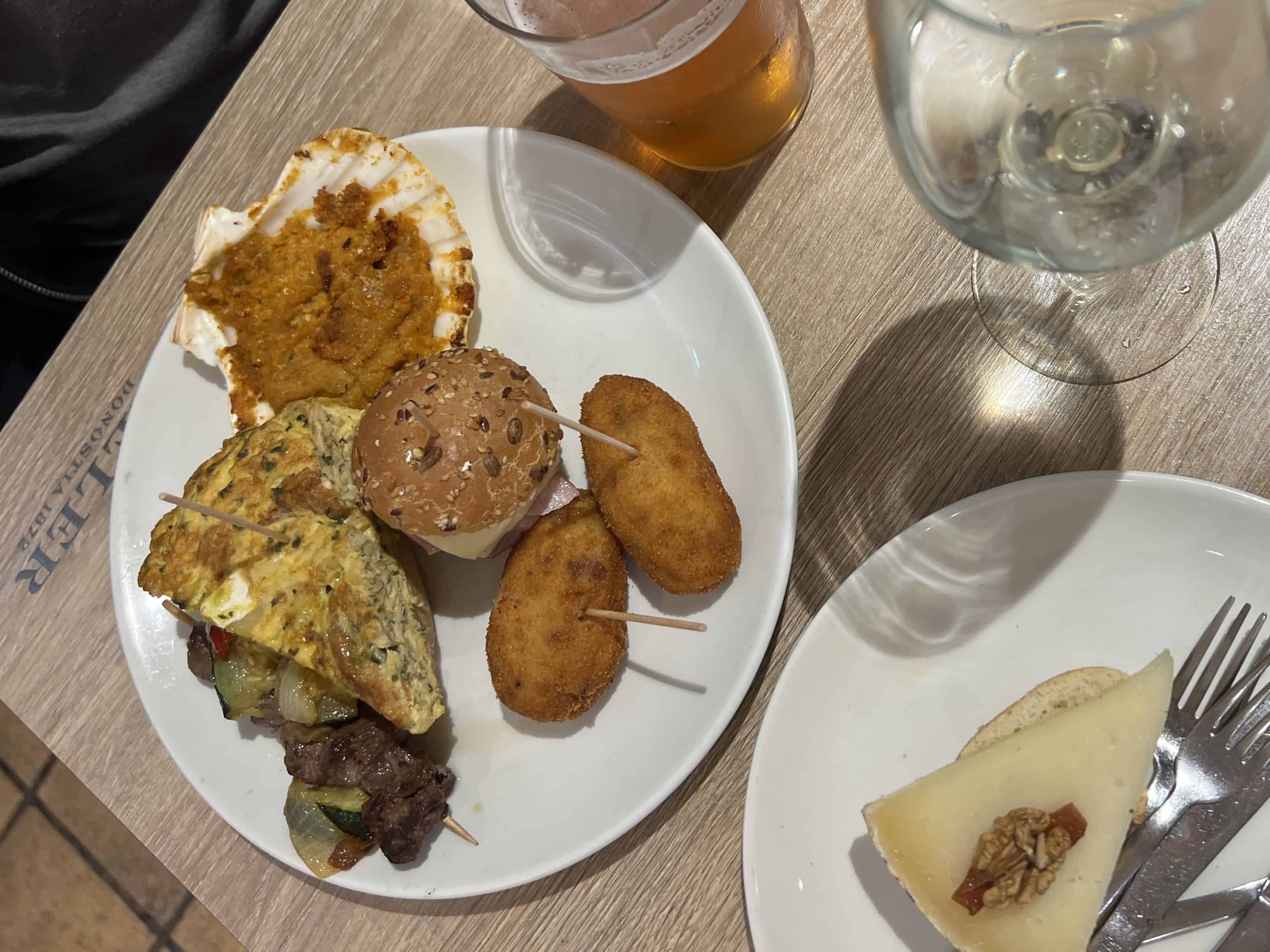 Overhead of two plates of popular pintxos on a wooden table next to a beer and water at a restaurant in San Sebastian.