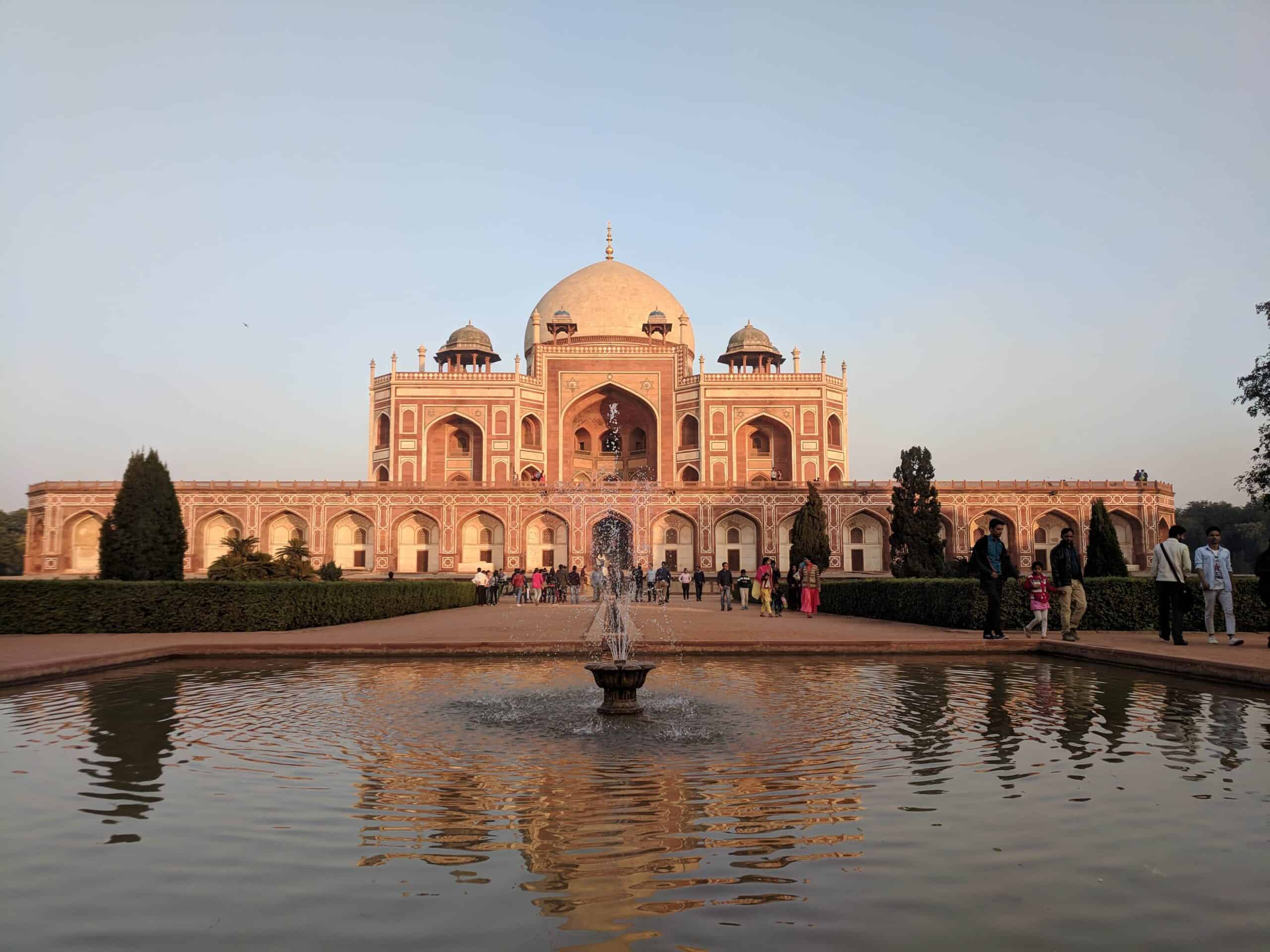 Visitors stroll around a reflective pool in front of the ornate Humayun's Tomb under a clear sky at sunset in Delhi, India.