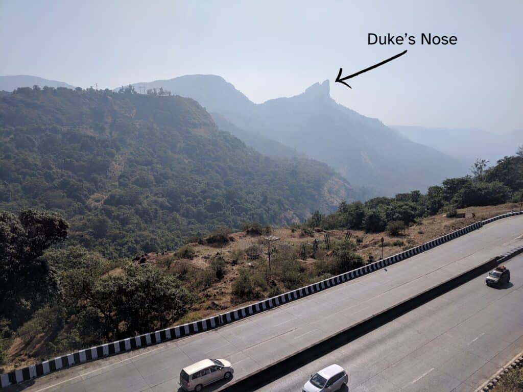 Faraway view of rocky hills in Lonavala with an arrow point to a ridge called Duke's Nose. A highway with a couple cars is in front of the hilly landscape.