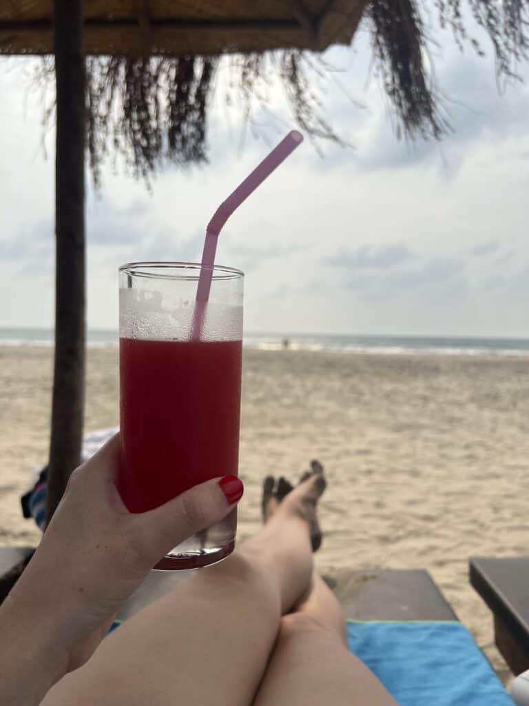 View of Ashvem beach with a girl holding a drink in front of the camera with her legs out on a beach lounger and a palm leaf umbrella above.