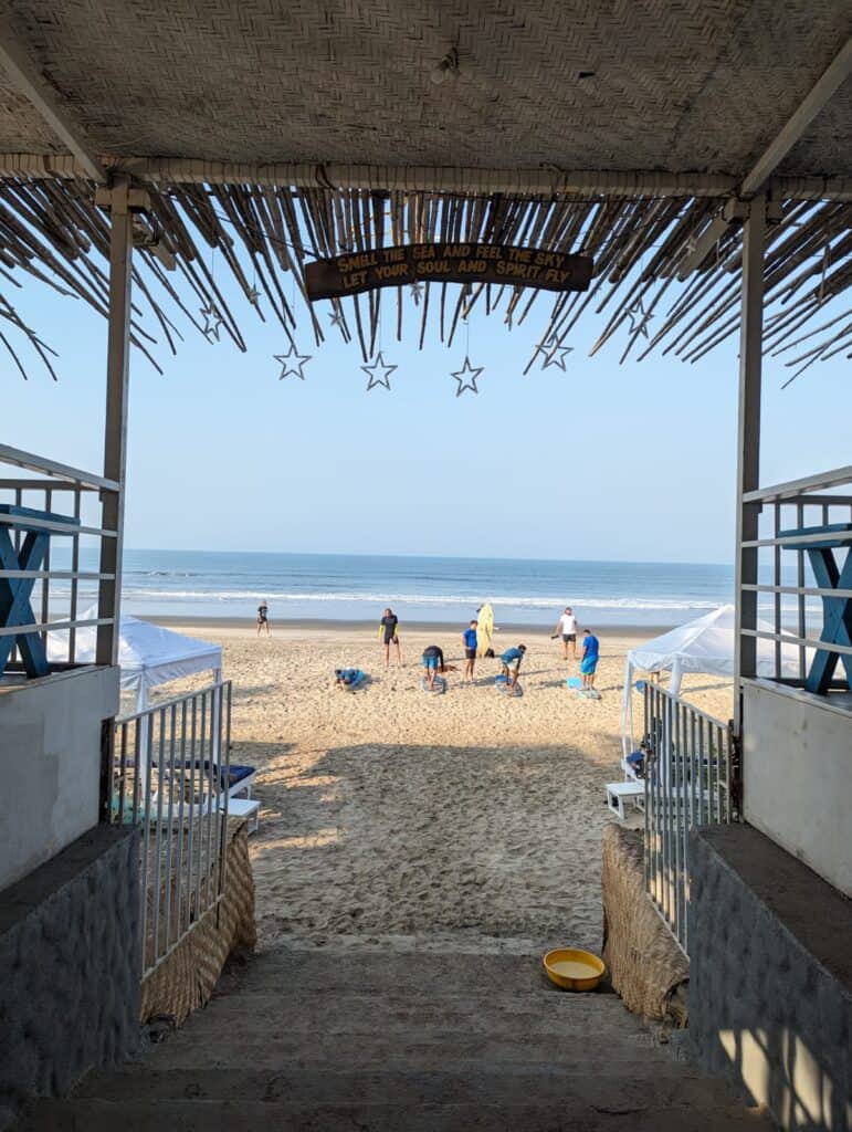 View of Morjim Beach from inside a shack with surfers practicing on the sand and the blue sky and ocean in the background.