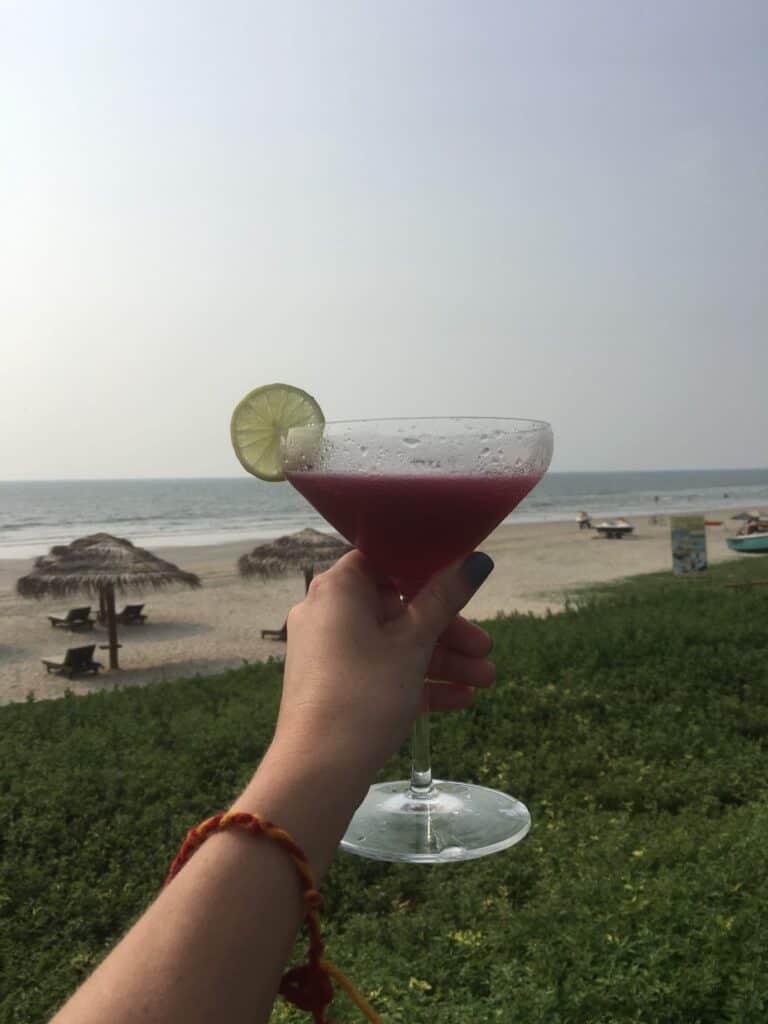 Close up of a cocktail held out front of the camera with the beach and ocean in the background at Taj Exotica in Goa.