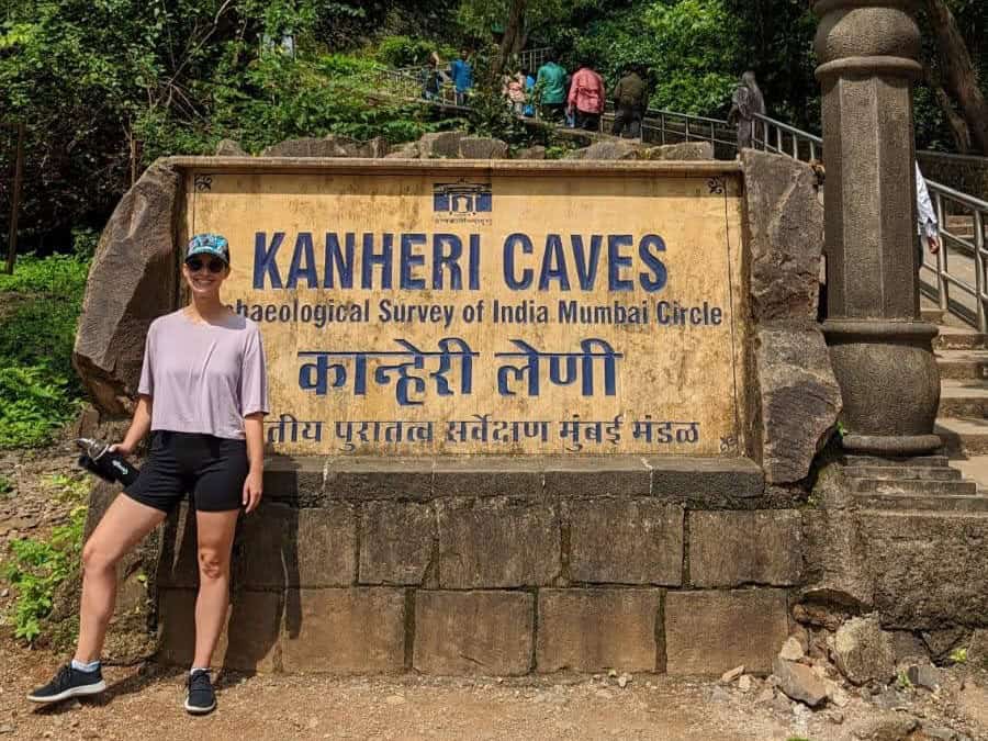 A girl dressed for a hike with a hat and waterbottle standing in front of the sign for the Kanheri Caves at Sanjay Gandhi National Park in Mumbai