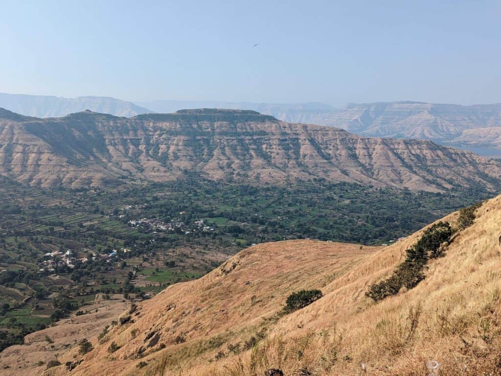 A panoramic view of the Sahyadri mountain range with its layered hills and valleys. In the foreground, golden-brown grass covers rolling hills, leading to a patchwork of green fields and small clusters of houses in the middle ground. A blue/gray sky in the background.