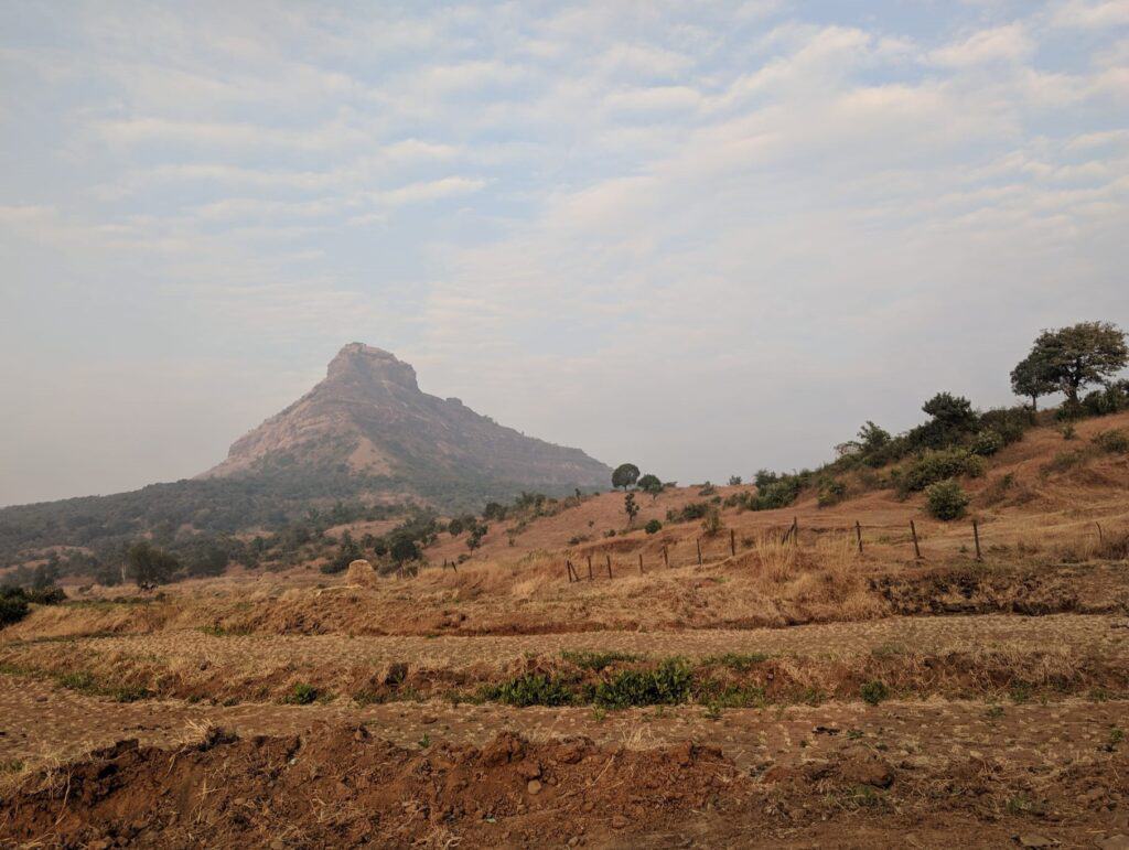 Brown dirt land with a small hill in the foreground and Tikona Fort raised up on a hill in the distance.