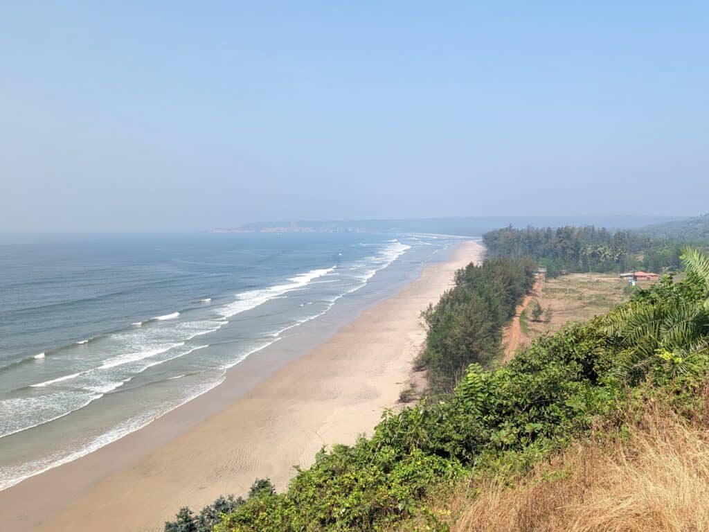 A panoramic view of Aare Ware beach with a long stretch of golden sand edged by gentle waves and backed by lush greenery under a clear blue sky.