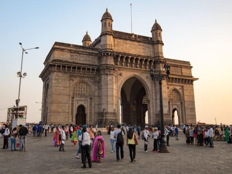 The Gateway of India in Mumbai, which is large stone landmark with 3 arches, with tourists wandering around in front of it and a blue sky background.