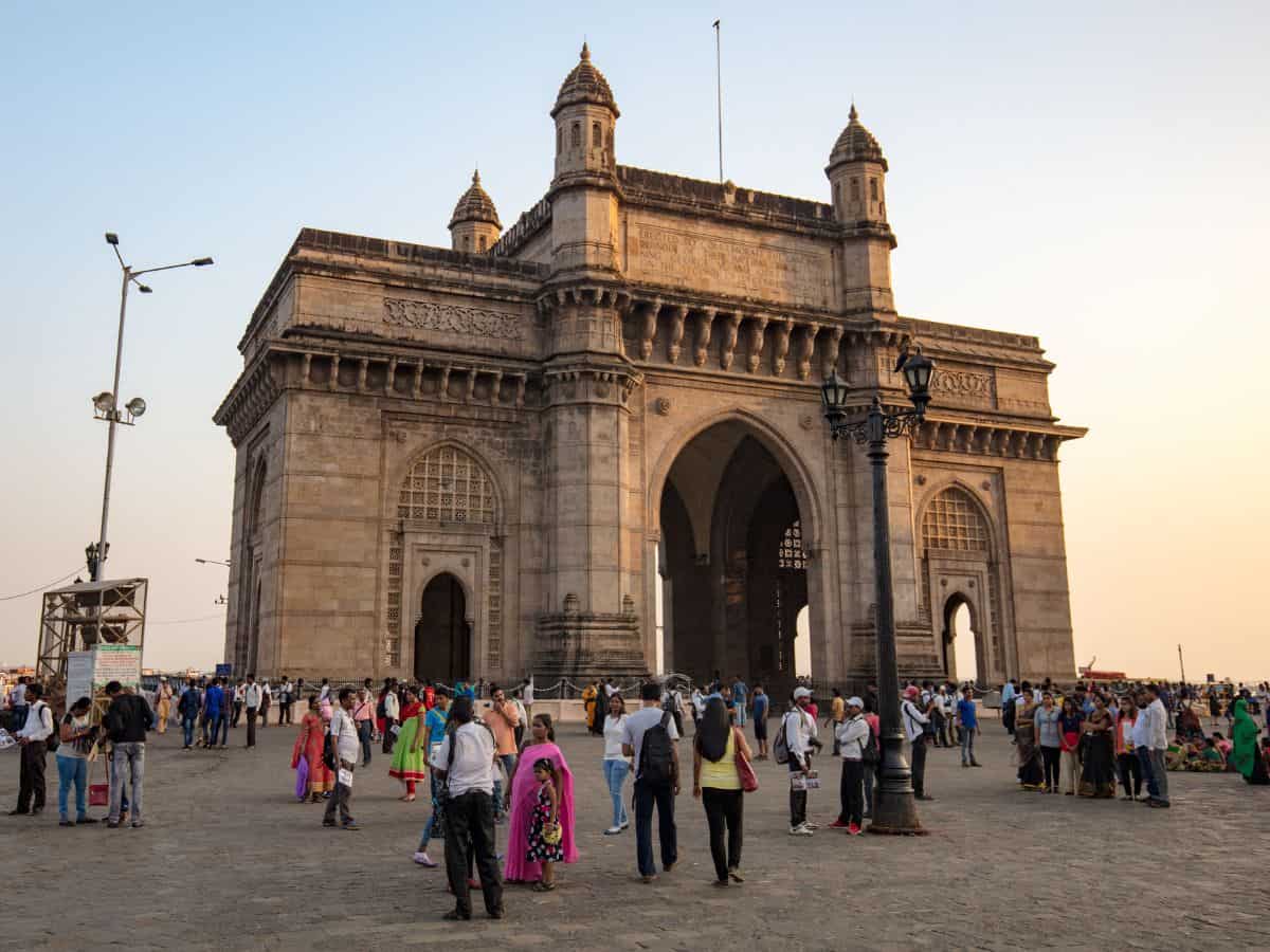 The Gateway of India in Mumbai, which is large stone landmark with 3 arches, with tourists wandering around in front of it and a blue sky background.