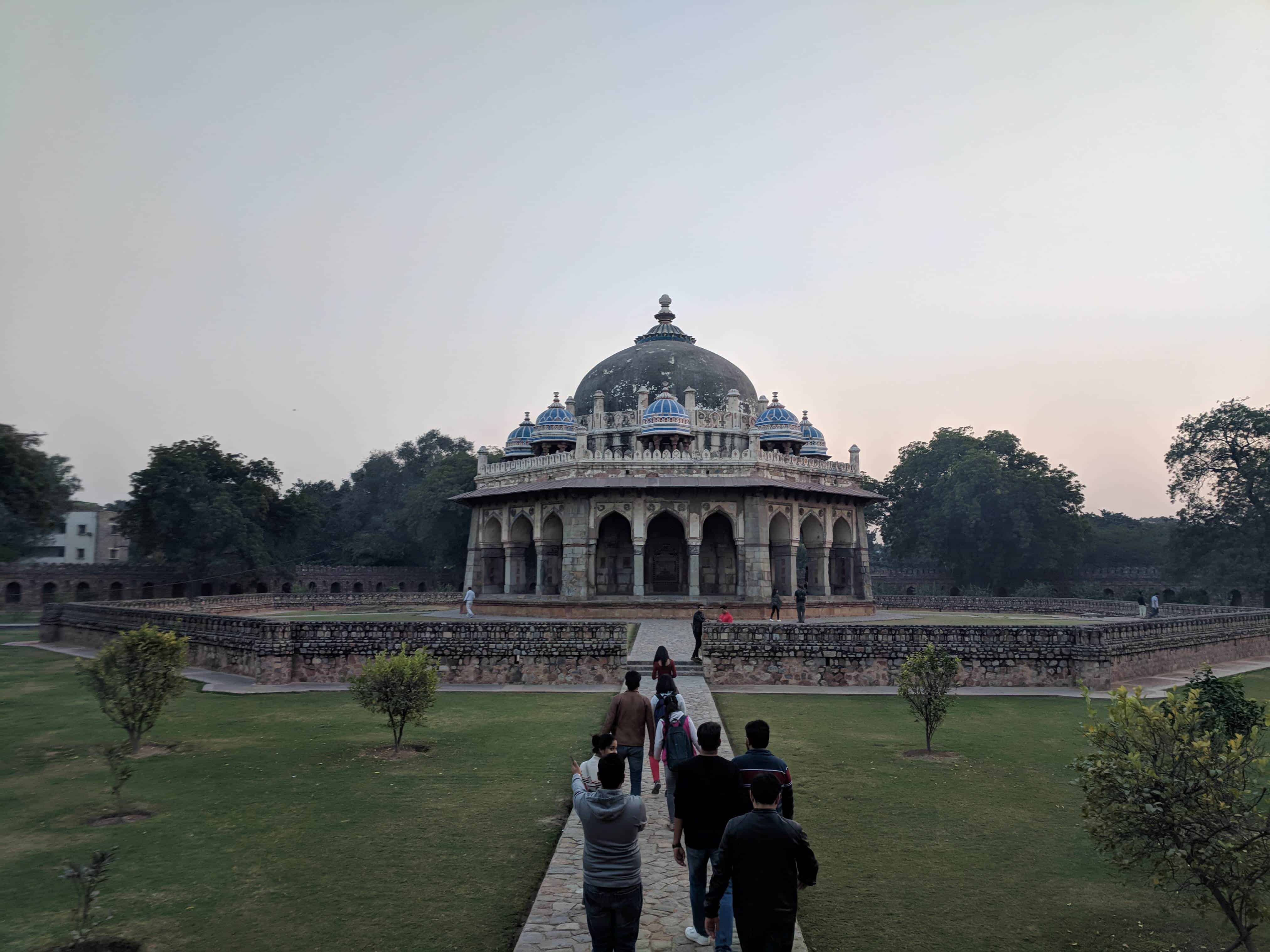 Visitors approaching the intricately detailed Isa Khan Niyazi's tomb located in the Humayun's Tomb complex in Delhi at dusk, with the structure featuring Mughal architecture surrounded by well-maintained gardens and walkways.