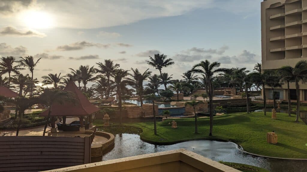 View from Juhu Beach Marriott hotel overlooking a luxurious pool area surrounded by lush palm trees, with a glimpse of the calm sea in the distance under a soft morning sky with the sun peeking through scattered clouds.