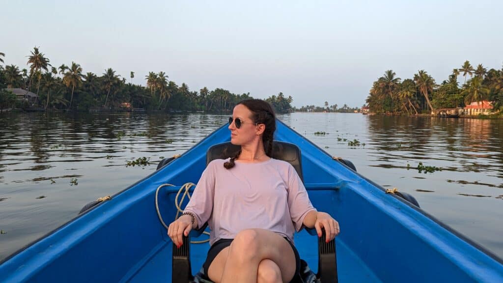 A woman sits at the stern of a blue rowboat on the calm backwaters in Kerala, surrounded by tropical palm trees at dusk.