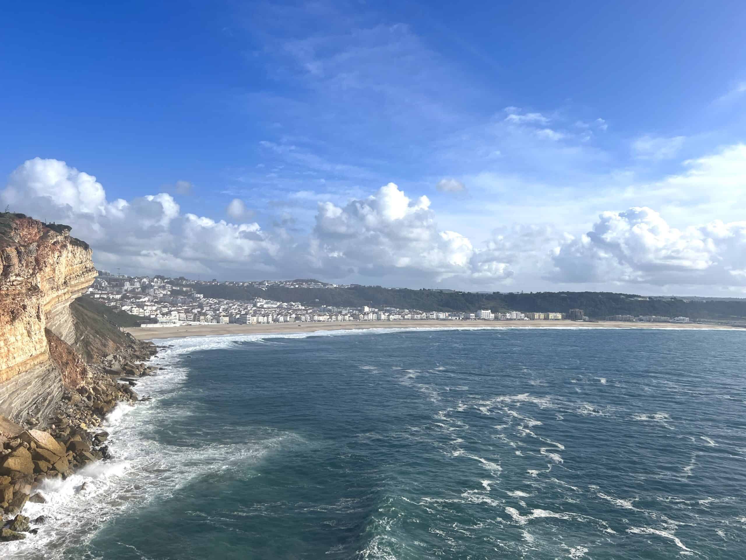 Cliffside view of Nazaré Beach in Portugal, showing the expansive sandy shore, waves in the Atlantic Ocean, and the town in the distance under a partly cloudy sky.