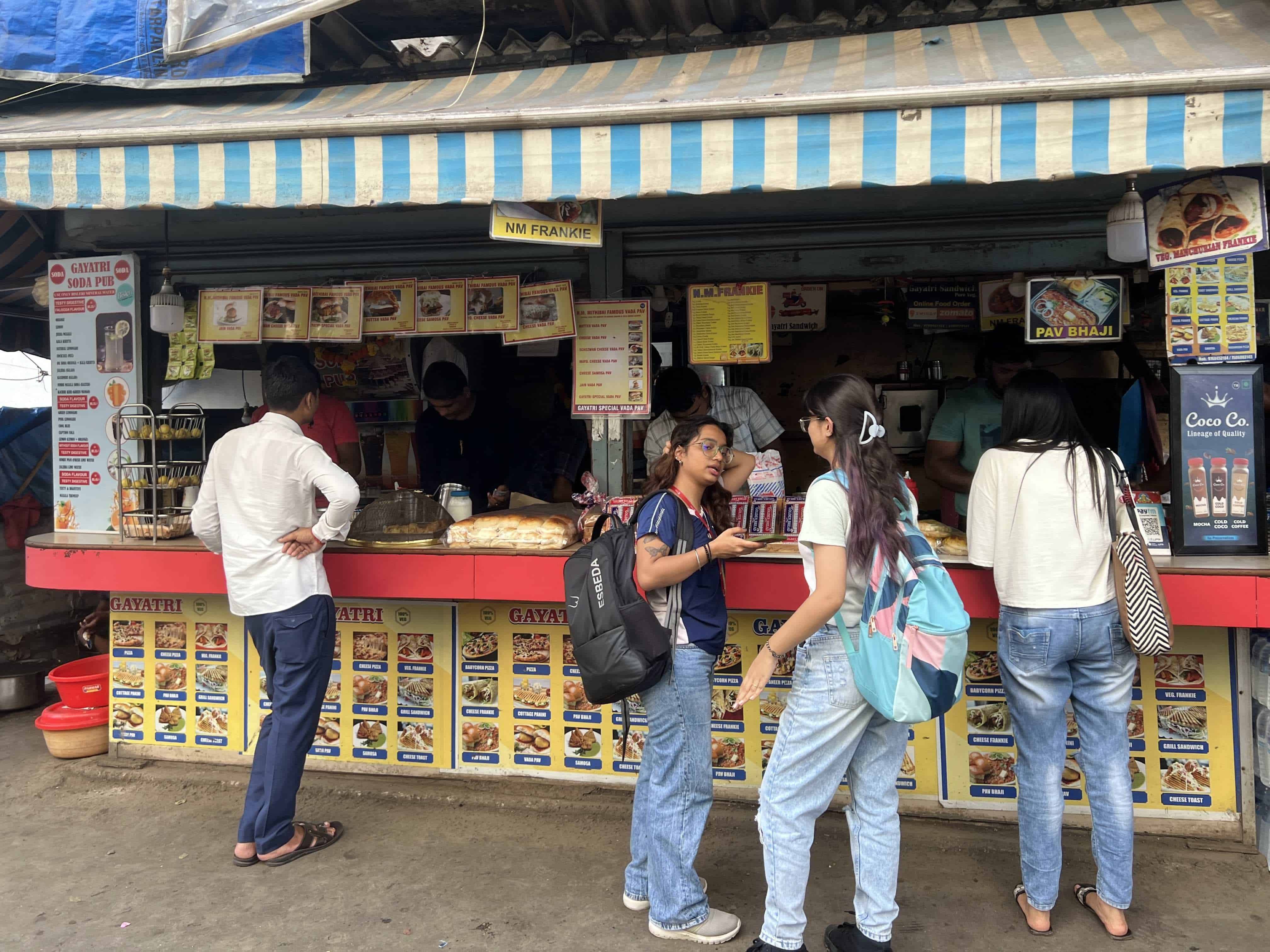 Customers at a street food stall with a blue and white striped awning in Mumbai, India. The stall displays a variety of food options on menus and serves popular Indian fast food like pav bhaji. People are seen ordering and waiting for their food.